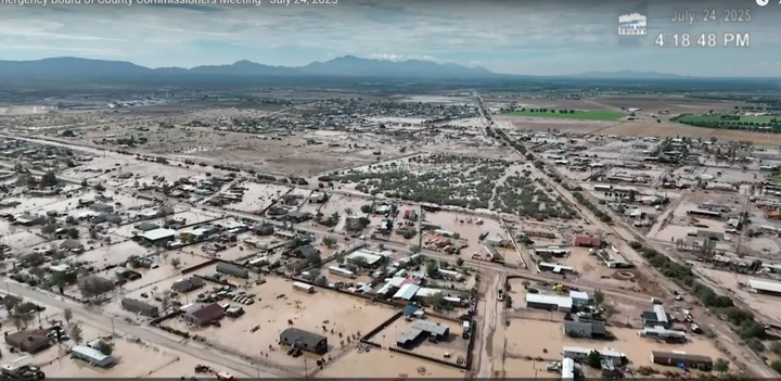 Aerial view of flooded neighborhoods with muddy streets and standing water, mountains in the distance — July 24, 2025