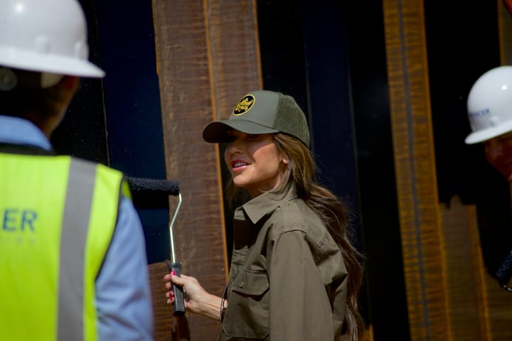 Kristi Noem stands in front of a newly painted section of the U.S.-Mexico border wall in Santa Teresa, accompanied by customs and border agents.