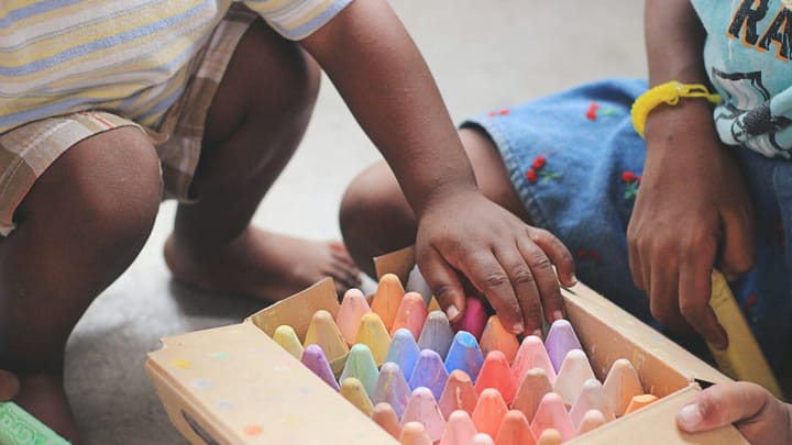 Two young children sit on the floor playing with a box of colorful sidewalk chalk, their hands reaching for pieces during an early learning or preschool activity.